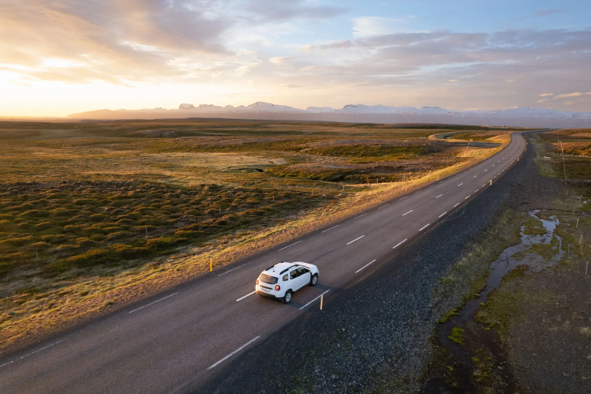 A white car driving on an empty road through a vast open landscape at sunset.